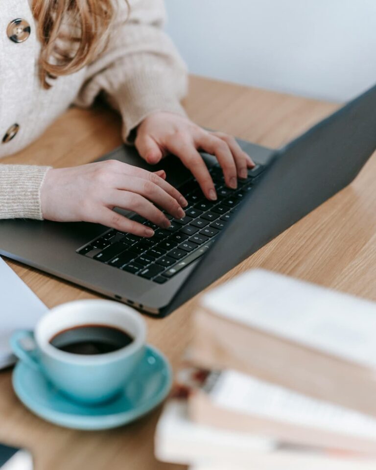 Alguien estudia en la laptop, con un café y una pila de libros sobre la mesa.