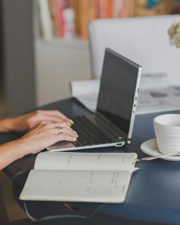 Manos escribiendo en una laptop, con un cuaderno abierto y una taza de café al lado.